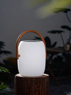 A Clérachic solar lantern with a matt white body on a log round, its handle in a wood-grain print, among blurred leaves.