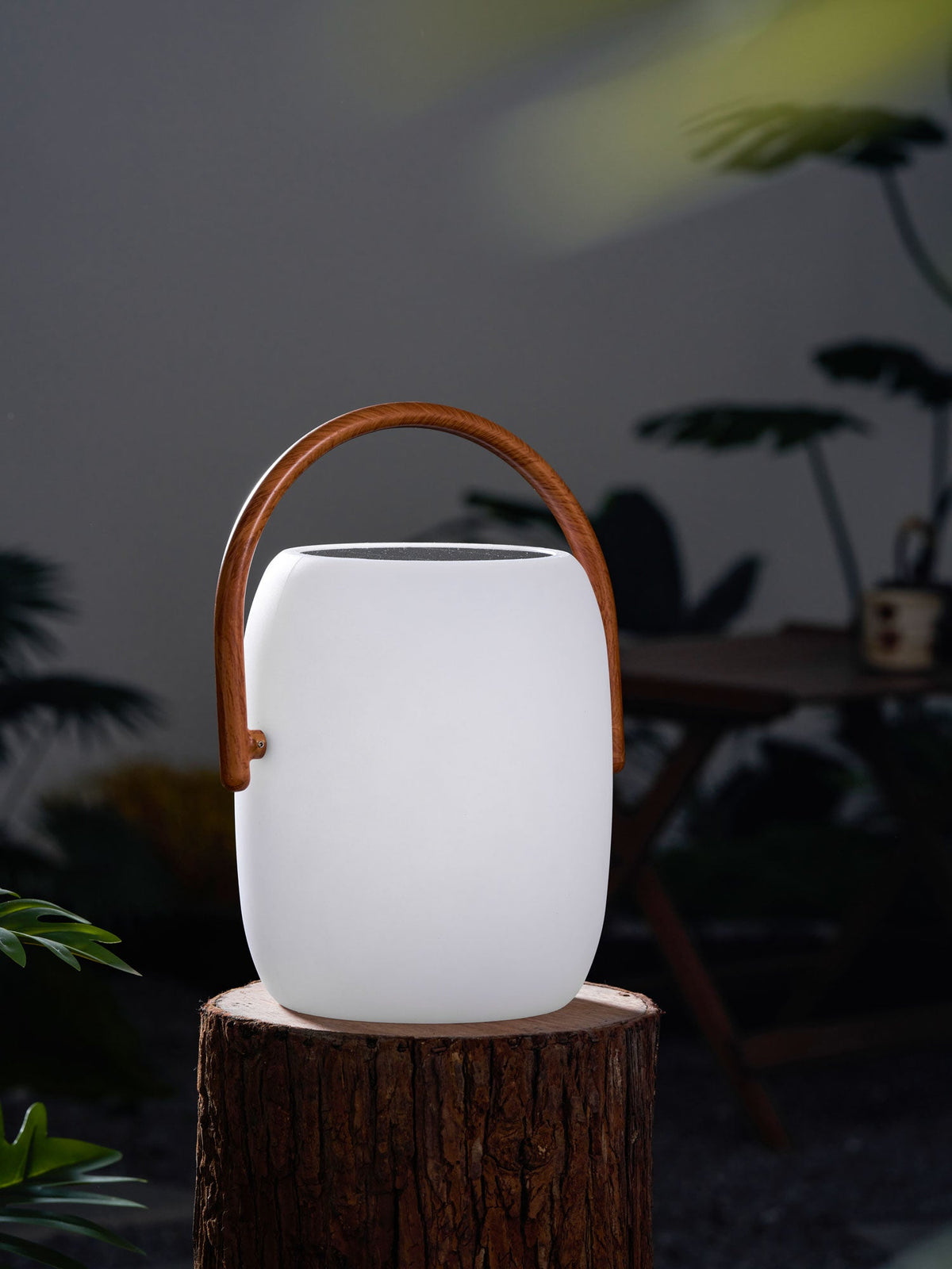 A Clérachic solar lantern with a matt white body on a log round, its handle in a wood-grain print, among blurred leaves.