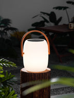 Gently glowing Clérachic lantern on a tree stump in a night garden, with green leaves in front and a table behind.