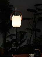 Small Clérachic lantern lit on a slim black pole in a dark garden, with a kettle and cup on a table at the bottom.