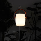 A Clérachic lantern glowing warm on a pole against the silhouettes of large dark leaves at night.