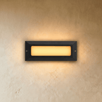 Clérachic small rectangular outdoor step light in dark grey with a frosted panel glowing warm white on a plaster wall.