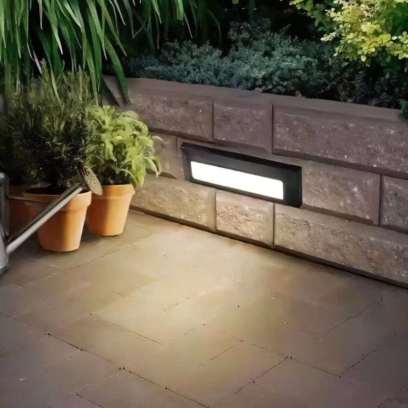 Clérachic dark rectangular step light set into a stone retaining wall beside terracotta pots, casting soft light across the paving.