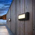 Row of Clérachic dark grey wall lights with warm glowing panels running along a weathered timber wall beside a lake at dusk.