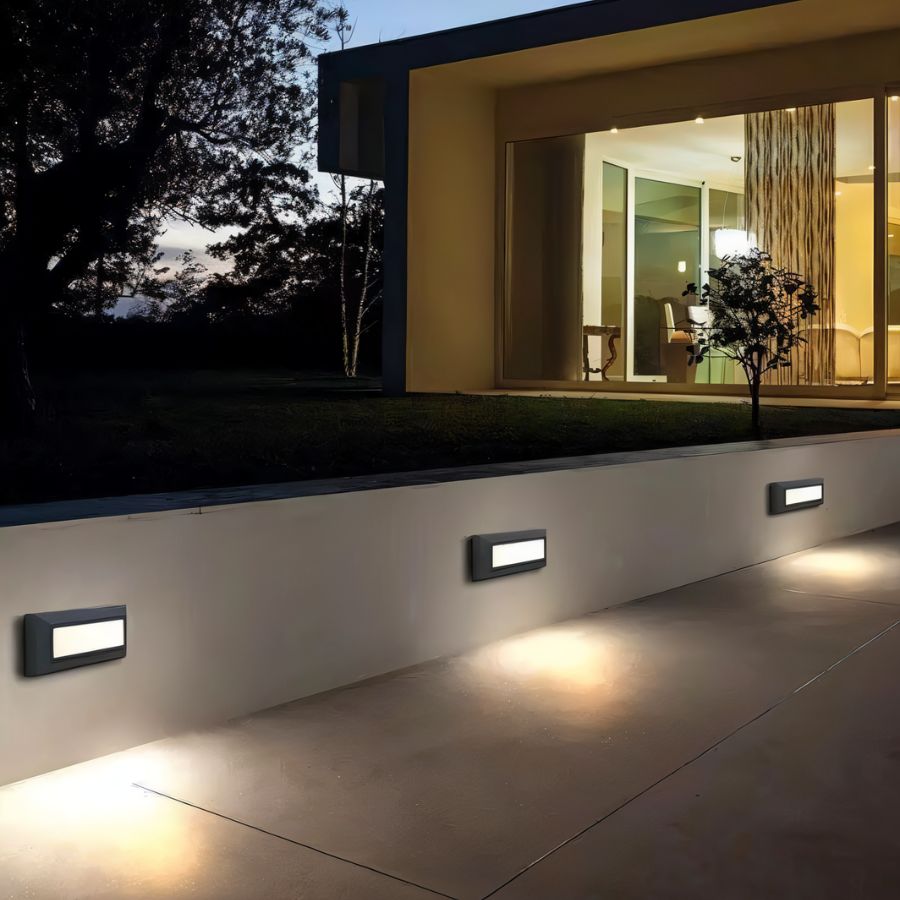 Clérachic dark grey step lights spaced along a rendered garden wall, throwing warm light onto the patio below at dusk.