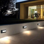Clérachic dark grey step lights spaced along a rendered garden wall, throwing warm light onto the patio below at dusk.