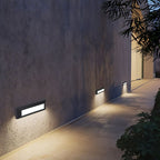 Clérachic dark grey wall lights along a rough concrete wall, washing the path beside it with warm light at night.