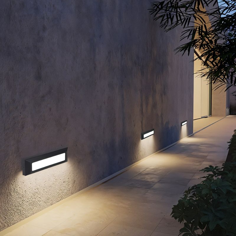 Clérachic dark grey wall lights along a rough concrete wall, washing the path beside it with warm light at night.