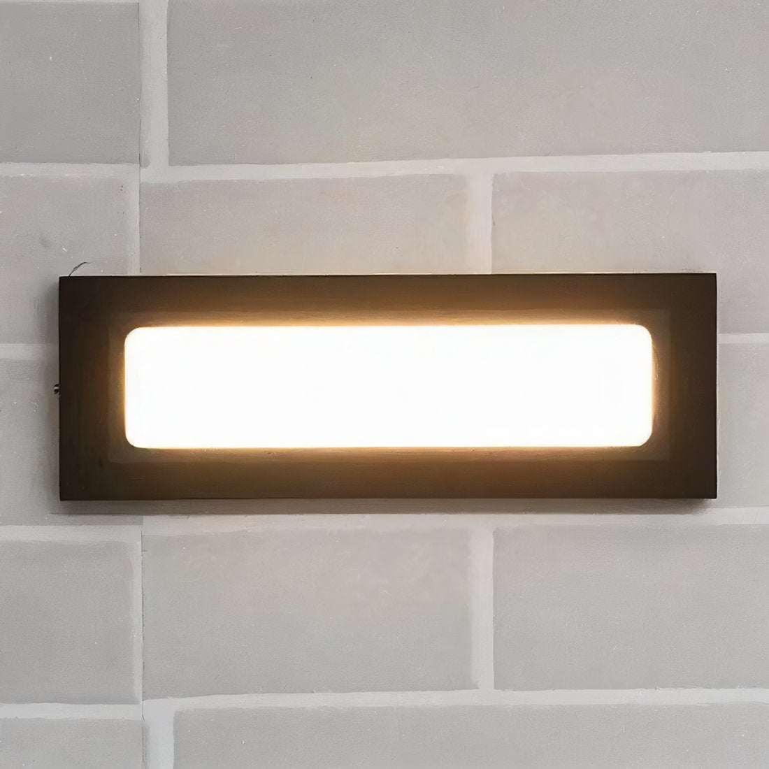 Close view of a Clérachic dark rectangular wall light with a bright warm glowing panel mounted on pale grey brick.
