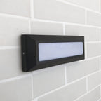 Angled view of a Clérachic dark grey wall light with an unlit frosted panel and bevelled frame on pale grey brickwork.