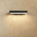 Clérachic black aluminium bar wall light, short length, glowing warm white along its underside against a limewashed plaster wall.