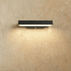 Clérachic black aluminium bar wall light, short length, glowing warm white along its underside against a limewashed plaster wall.
