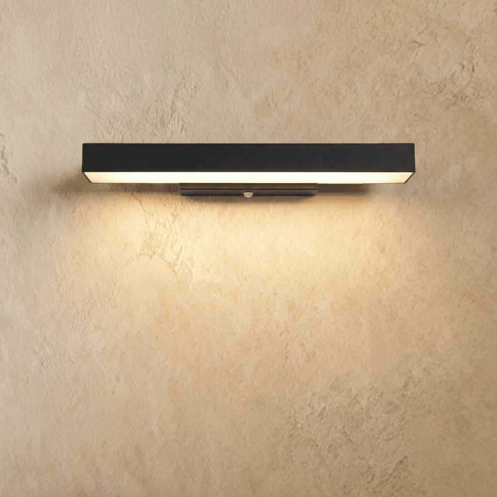 Clérachic long black aluminium bar wall light with a warm white glow washing down a textured limewashed wall, small sensor on the bracket.