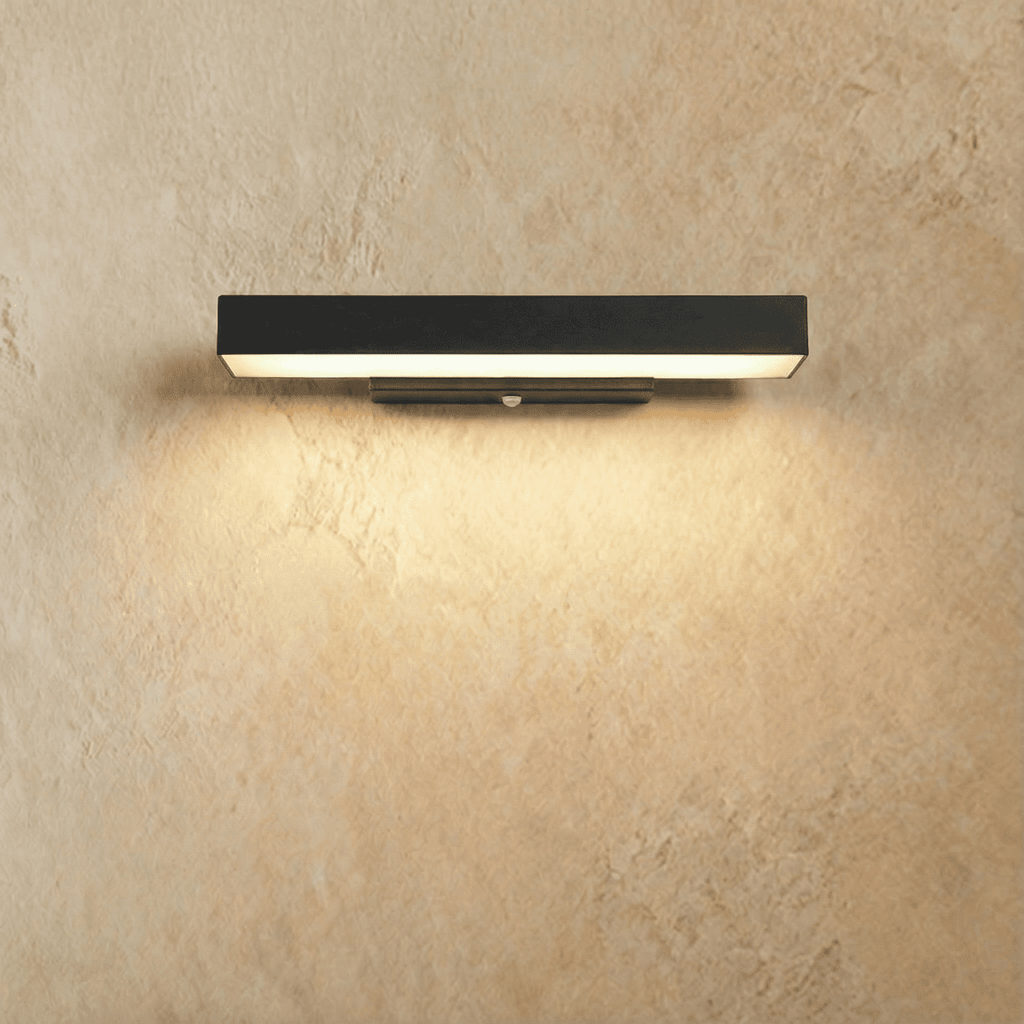 Clérachic mid-length black aluminium bar wall light lit warm white, with a slim wall bracket and sensor, on a limewashed wall.