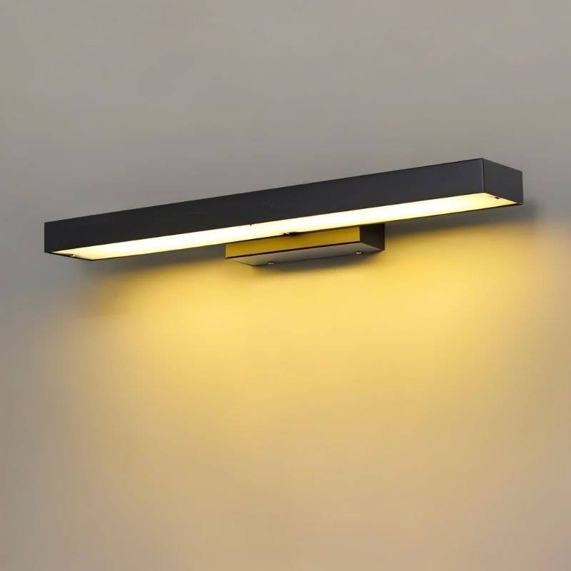 Clérachic long black bar wall light with a warm amber glow on a plain grey wall, seen at an angle with its brass-toned bracket.