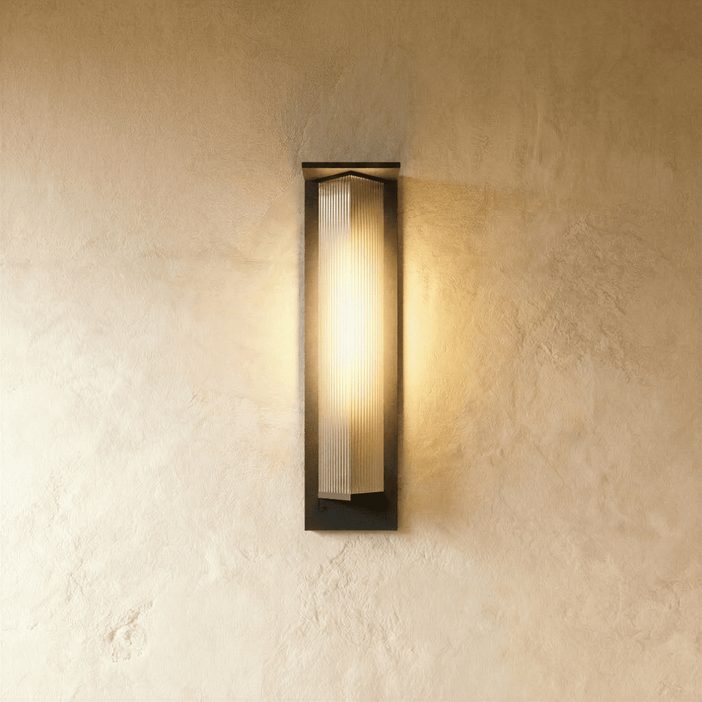 Clérachic outdoor wall light with a black metal frame and ribbed glass shade, glowing warm on a limewashed plaster wall
