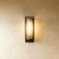 Clérachic outdoor wall light in the shorter height, a black metal frame around a ribbed glass shade, lit warm on plaster