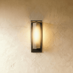 Clérachic outdoor wall light in the shorter height, a black metal frame around a ribbed glass shade, lit warm on plaster