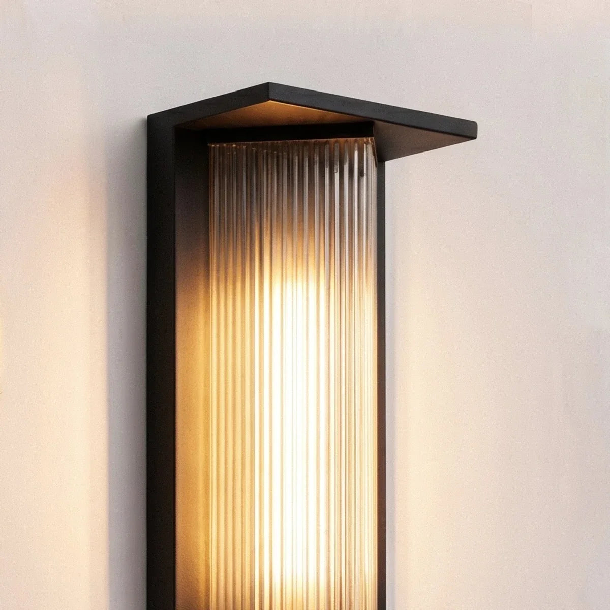 Close view of the Clérachic outdoor wall light: black metal hood and frame over a ribbed glass shade glowing warm