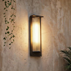 Clérachic outdoor wall light with a dark frame and ribbed glass shade, lit warm on a rough plaster wall beside trailing ivy