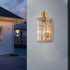 Lit Clérachic brass lantern wall light on a pale rendered wall at dusk, with a white timber house and string lights behind.