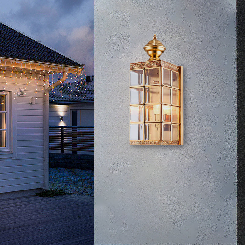 Lit Clérachic brass lantern wall light on a pale rendered wall at dusk, with a white timber house and string lights behind.