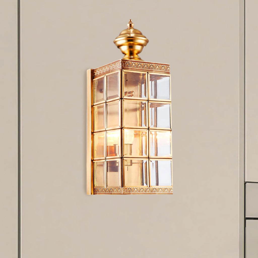 Clérachic brass and glass wall light, lit, on a warm grey wall beside a door frame, its etched brass band under the finial.