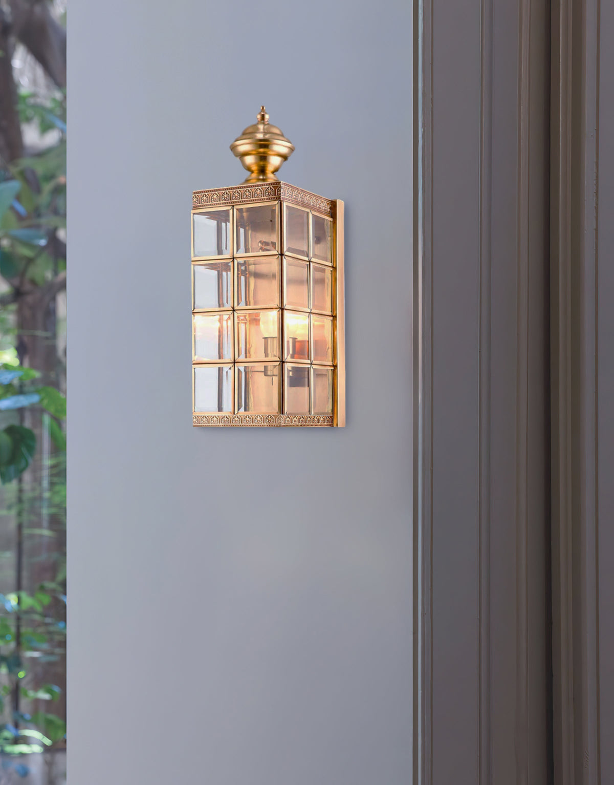 A Clérachic brass wall light, lit, on a pale blue-grey wall beside a dark door frame and green foliage.