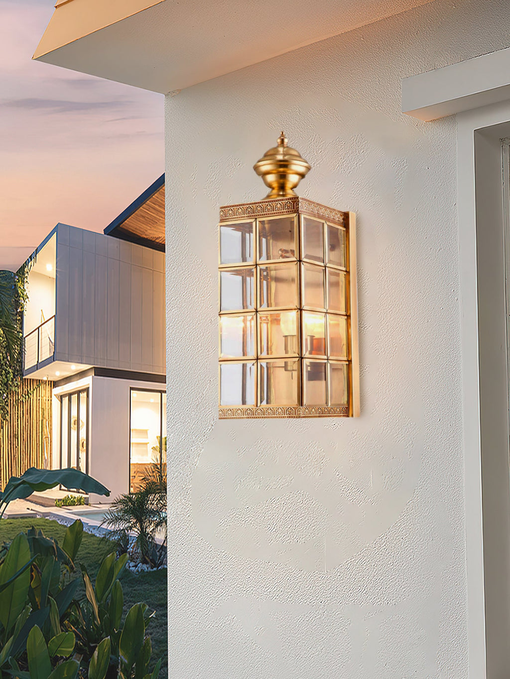Brass lantern wall light from Clérachic glowing on a white rendered wall at sunset, with a modern house and garden beyond.