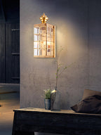 Clérachic brass wall light on a rough grey concrete wall, lit above a dark wooden bench with a vase of twigs and a cushion.