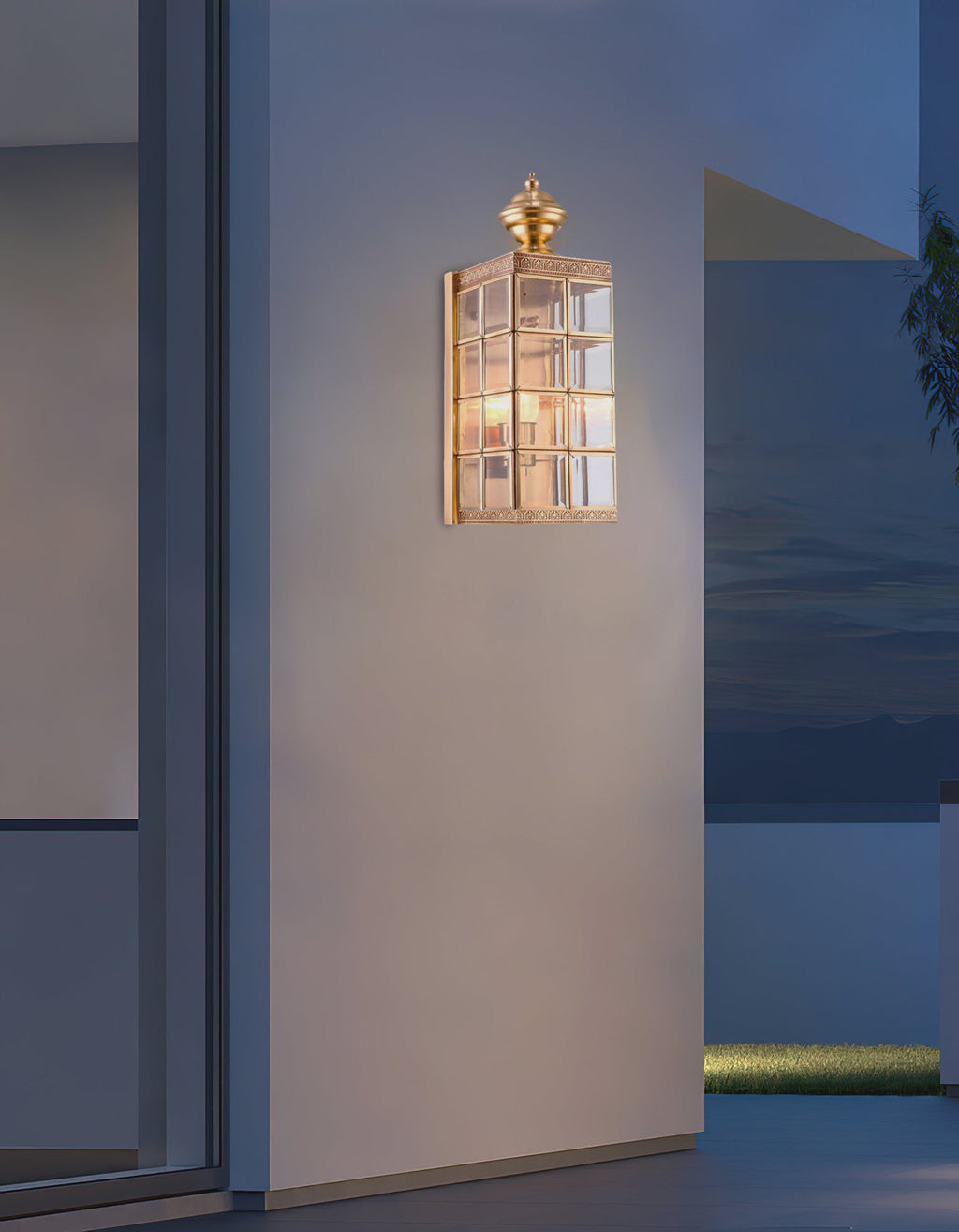 Lit Clérachic brass and glass wall light on a pale wall at dusk, beside a glazed door and a view across the water.
