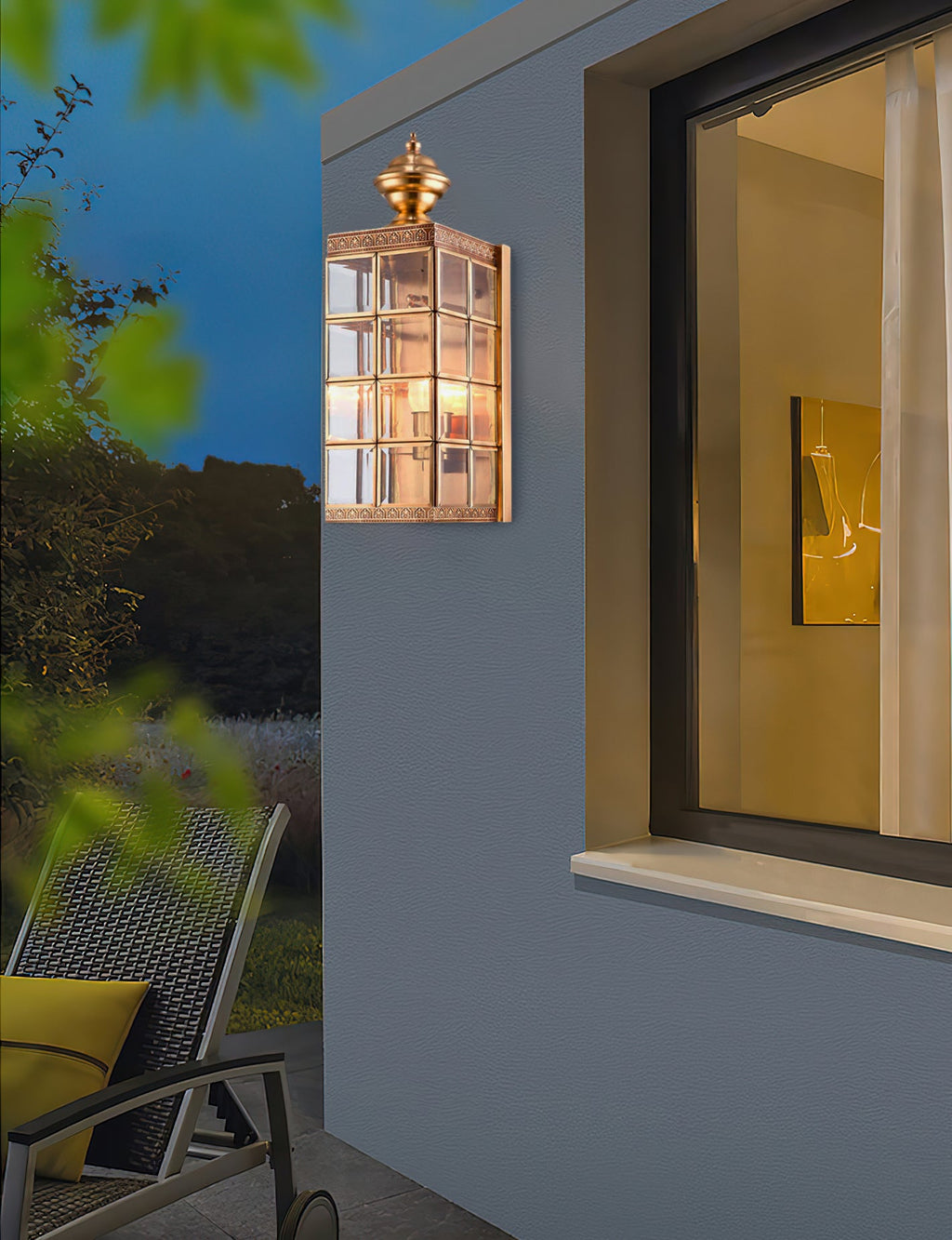 A Clérachic brass lantern wall light, lit, beside a dark-framed window at dusk, with a lounge chair on the terrace below.