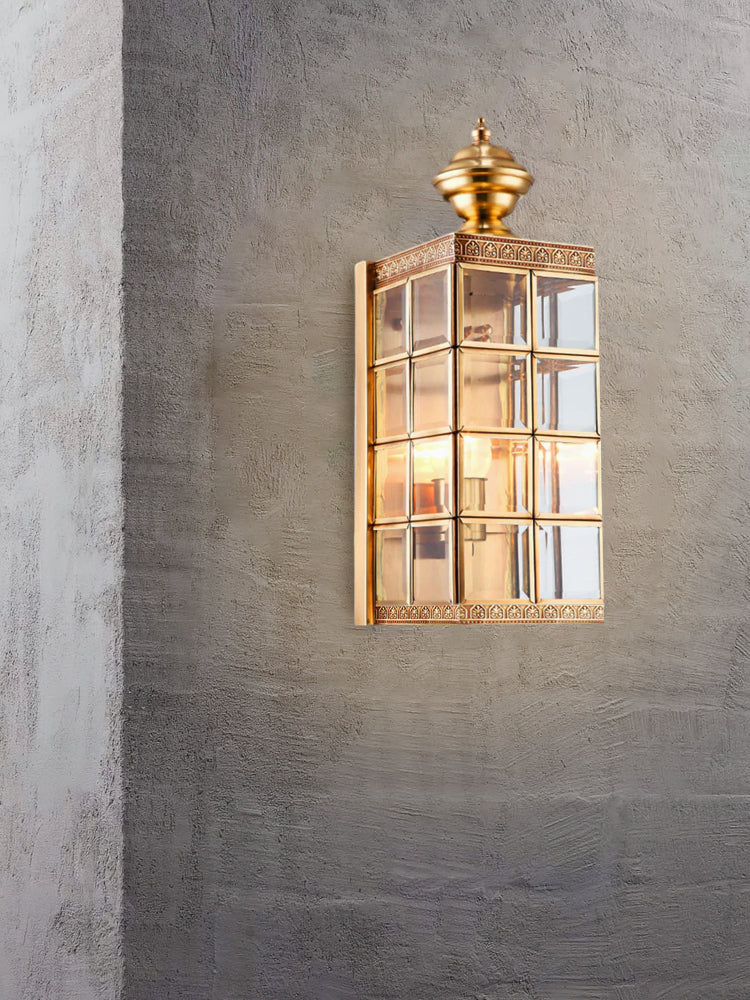 Clérachic brass wall light lit on a textured concrete wall, its gridded glass panes showing the bulb inside.