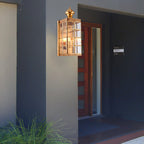 A Clérachic brass wall light, lit, beside a dark entrance with a red timber door, a house number plate and a clump of grass.