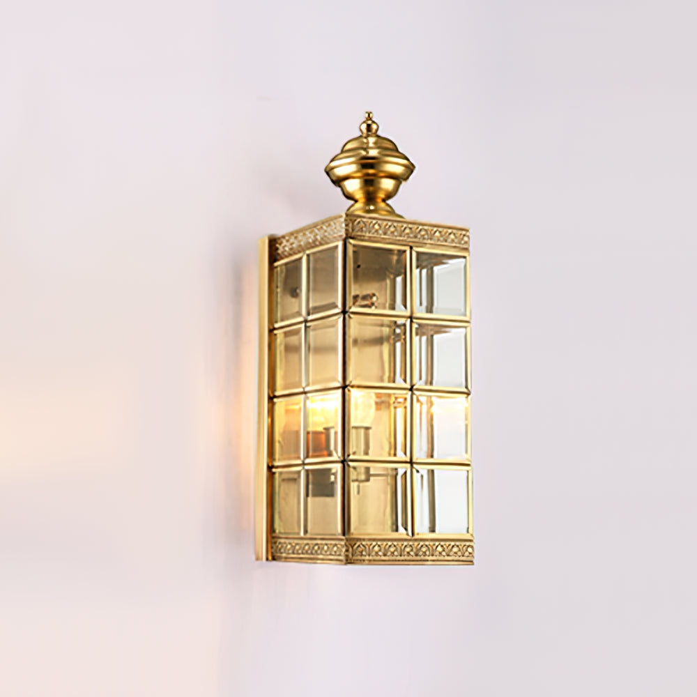 Lit Clérachic brass lantern wall light on a pale lilac wall, its glass panes glowing warm under the brass finial.