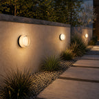Three round white-glass Clérachic wall lights along a rendered garden wall at dusk, beside a gravel bed and stone path.