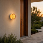 Round amber ribbed-glass Clérachic wall light glowing beside a tall wooden door on a pale rendered wall at sunset.
