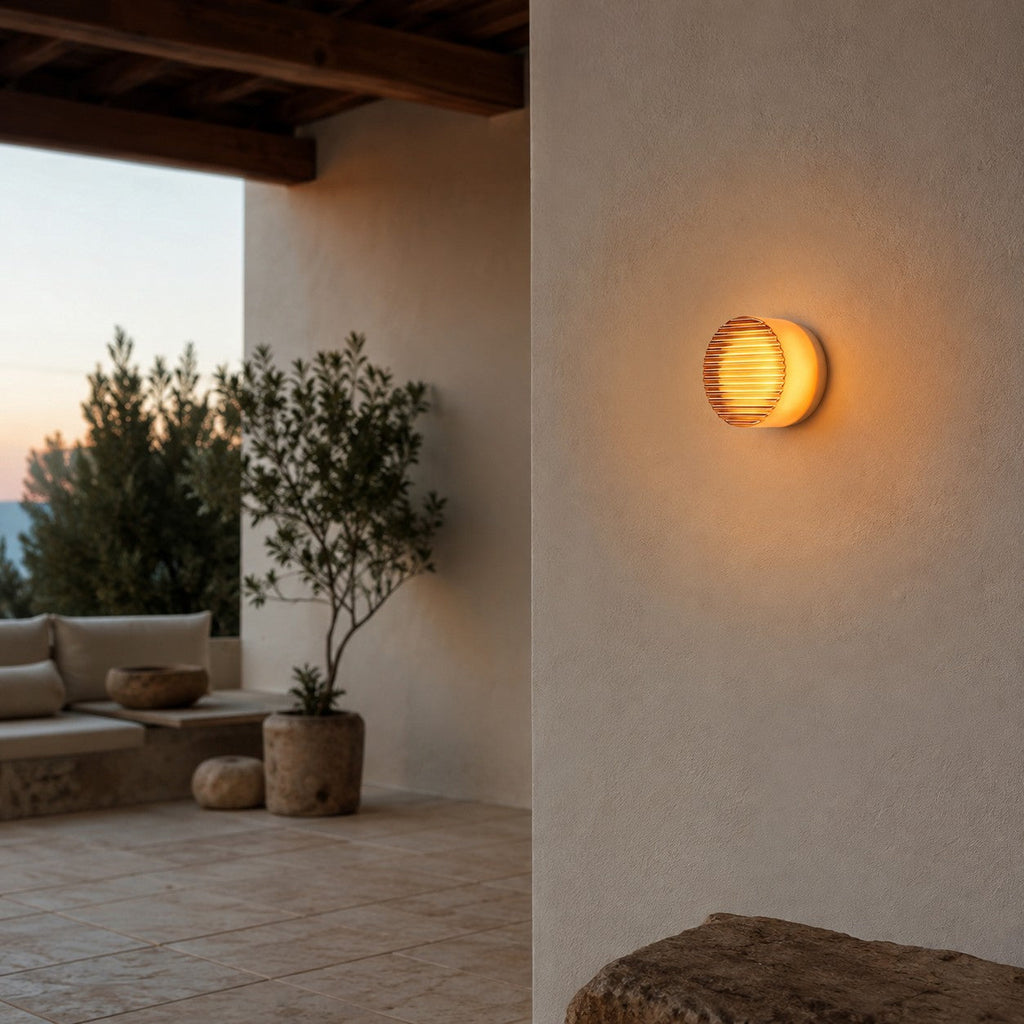 Amber ribbed-glass Clérachic wall light on a rendered wall under a timber-beamed porch, with a lounge corner and olive tree.