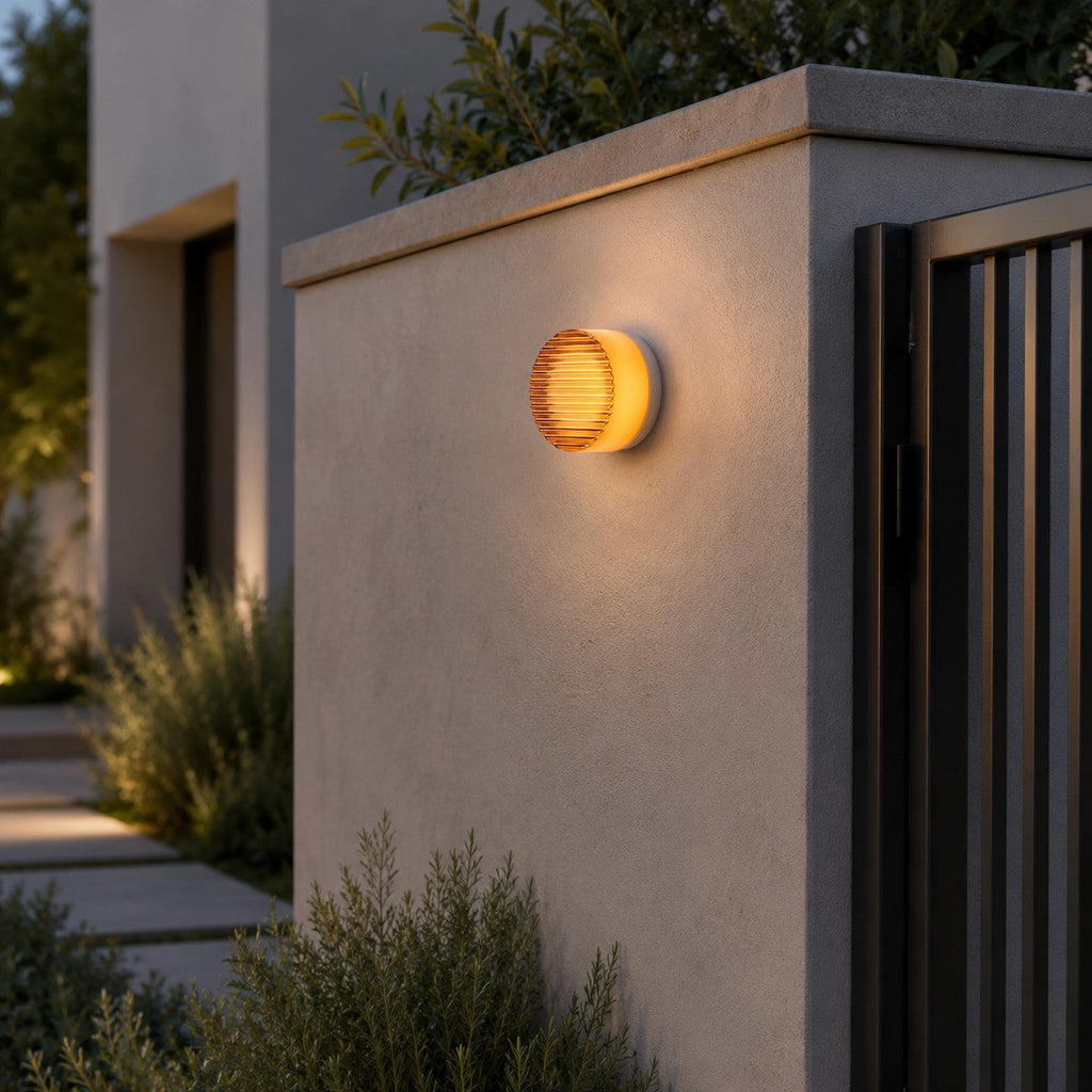 Amber Clérachic wall light on a gate-side rendered wall, glowing warm above a planting bed and a dark slatted gate.