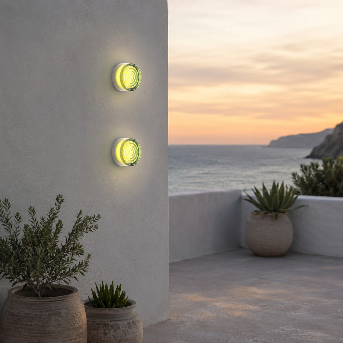 Two green-glass Clérachic wall lights stacked on a curved wall, with a sea view, potted olive and agave at sunset.