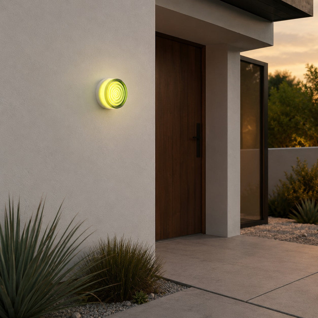 Green-glass Clérachic wall light beside a wooden entrance door at dusk, with grasses and gravel at the foot of the wall.
