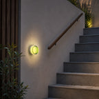 Green Clérachic wall light lighting a rendered stairwell wall beside a timber handrail and concrete steps at dusk.