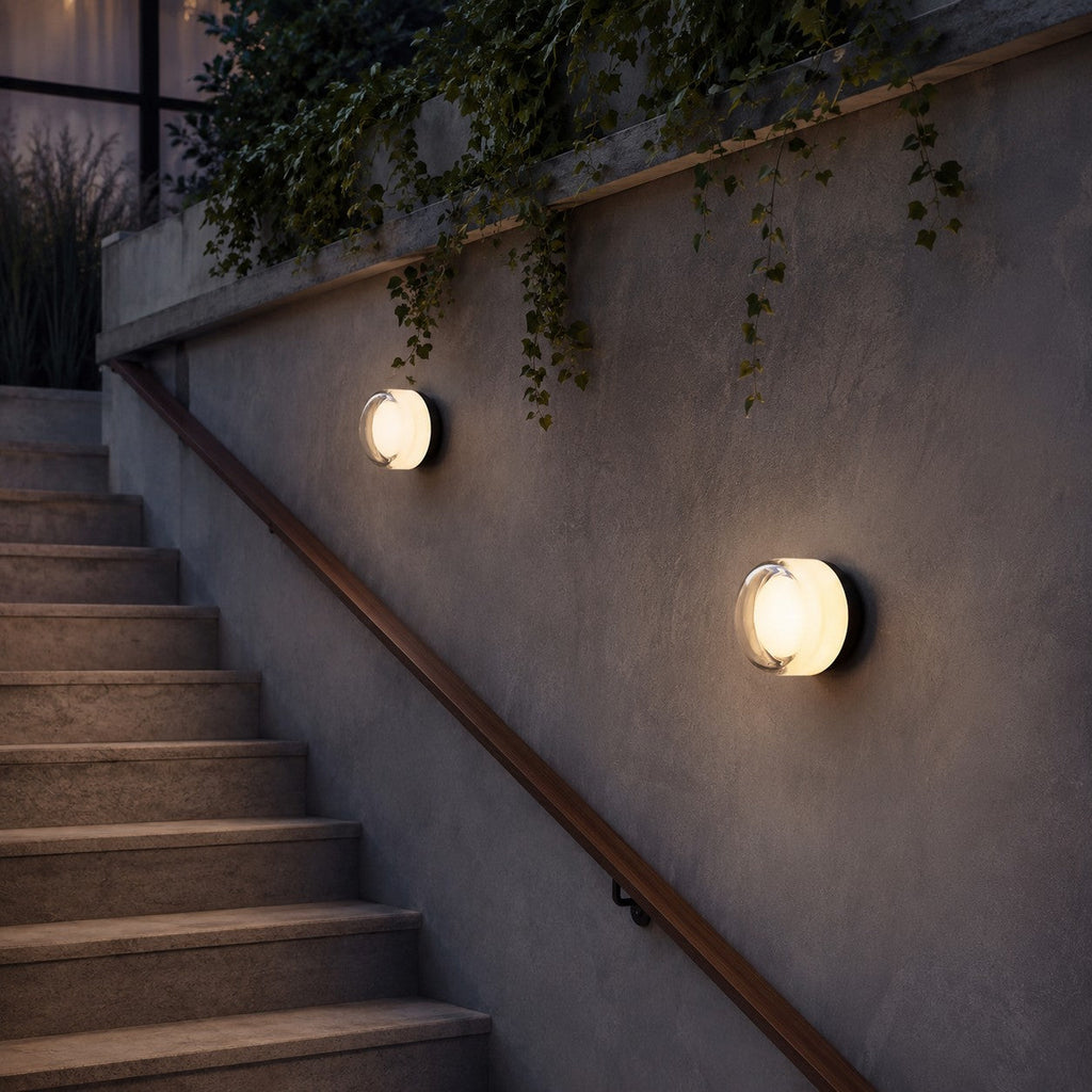 Two white-glass Clérachic wall lights on a stair wall, one small and one larger, beside a wooden handrail and trailing ivy.