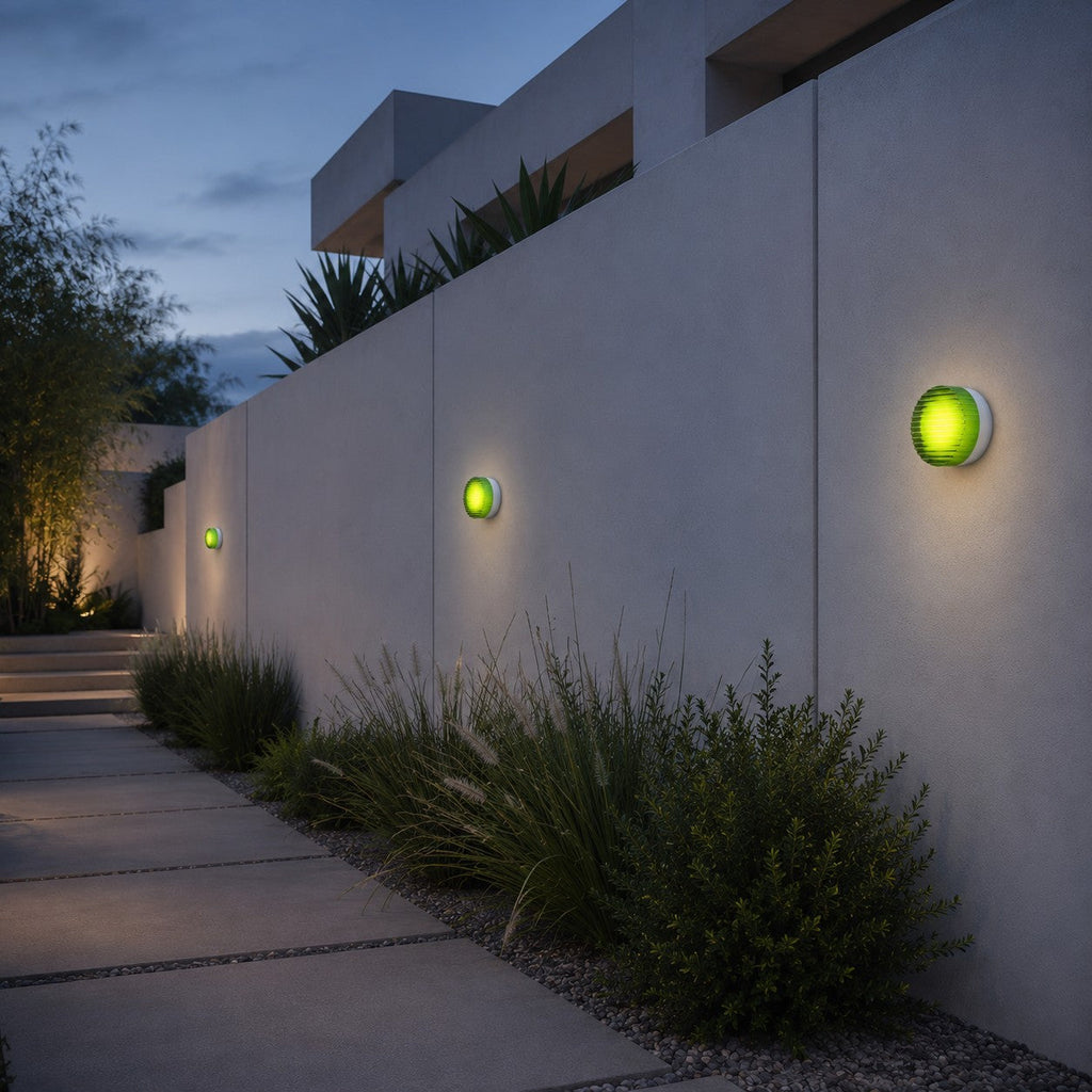 Green-glass Clérachic wall lights spaced along a long garden wall at blue hour, above clipped shrubs and a stone path.