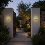 Green Clérachic wall lights on two tall wall piers flanking a stepping-stone path, with an olive tree lit behind.