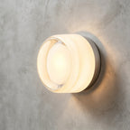 White frosted-glass Clérachic wall light with a clear lens at the centre, lit warm against a grey concrete wall.