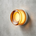 Amber glass version of this Clérachic wall light with a clear lens at the centre, glowing on a grey concrete wall.