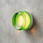 Green glass version of this Clérachic wall light with a clear central lens, casting a soft glow on a concrete wall.