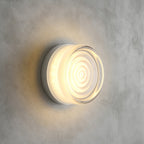 Rippled-ring clear glass Clérachic wall light lit warm white, mounted on a grey concrete wall with a soft halo.