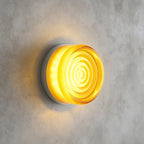 Yellow glass Clérachic wall light with concentric ripples on its face, lit and mounted on a grey concrete wall.
